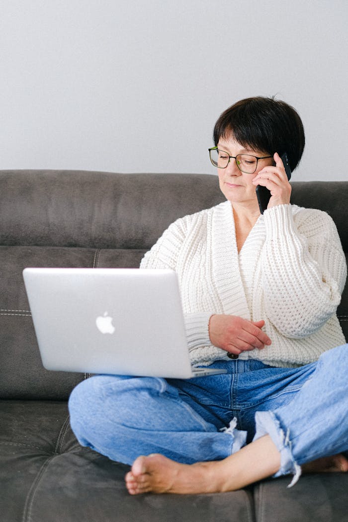 Elderly woman sitting on sofa multitasking with a laptop and phone.